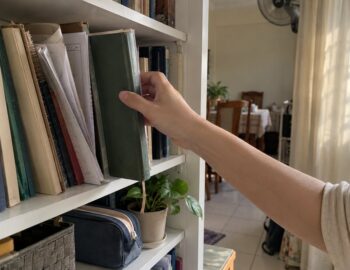 A hand pulls a hardback exercise book from an uneven shelf of school books, with a folded worksheet and pencil case beside it in soft window light.